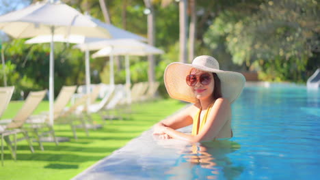 Una-Mujer-Bonita-De-Pie-En-Una-Piscina-Con-Un-Enorme-Sombrero-Blanco-Para-El-Sol-Y-Gafas-De-Sol-Sonríe-Mientras-Hace-Contacto-Visual-Con-La-Cámara