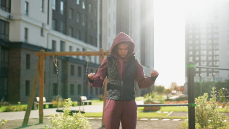 sports woman in hoodie and vest stretching arms outdoors in urban playground with autumn foliage and sunlight illuminating movement and fabric texture for warm up before intense workout session