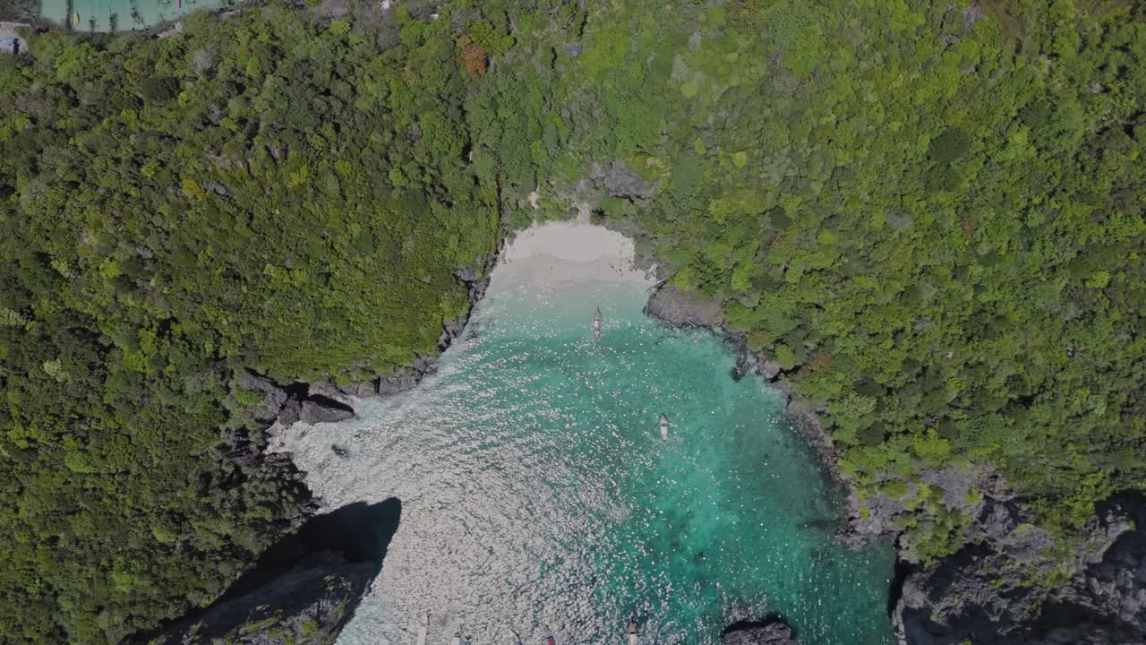Drone ascends vertically with a top-down perspective, revealing a turquoise lagoon framed by limestone cliffs and tropical forest in Phi Phi Islands, Thailand.