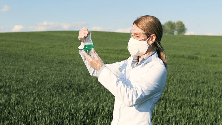 Caucasian researcher woman in white coat and goggles looking at test tube in the green field