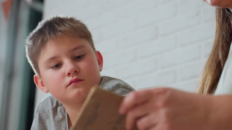 close view of hand flipping book page with boy in grey top attentively observing, focused serious expression, indoor cozy setting with soft daylight and white brick wall background