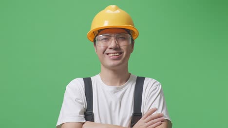 close up of asian man worker wearing goggles and safety helmet smiling and crossing his arms while standing in the green screen background studio