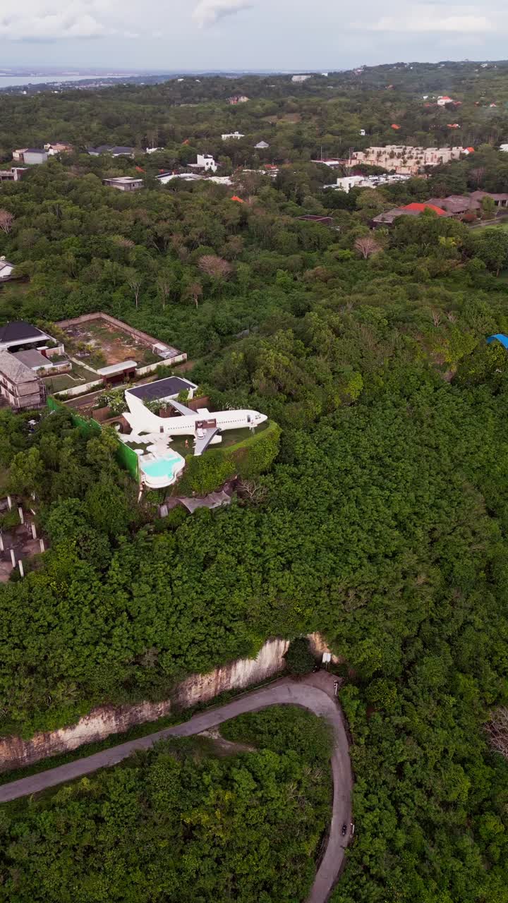 Vertical aerial shot shows a Bali airplane villa set on a hilltop with a wide infinity pool facing the ocean while paragliders move along the coastline giving a clear view of this unique luxury stay
