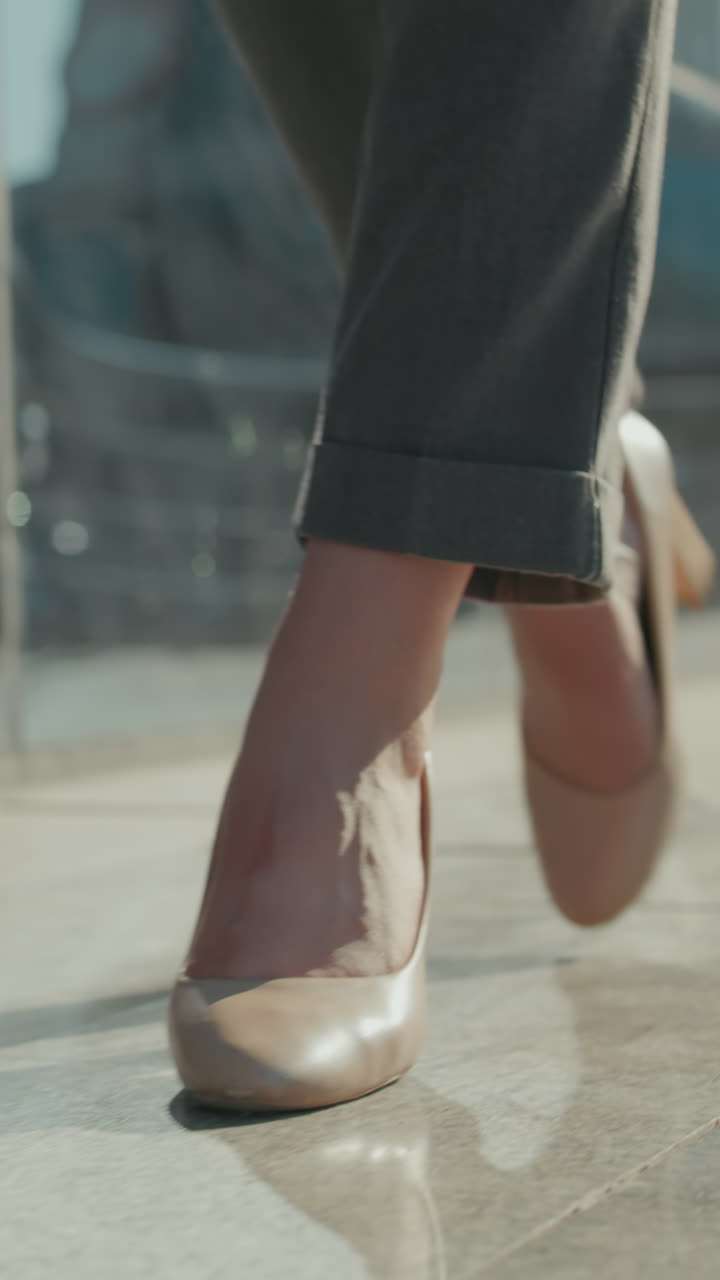 Lower angle view of girl walking confidently in high heels along modern urban walkway with glass building reflecting surroundings and soft bokeh light