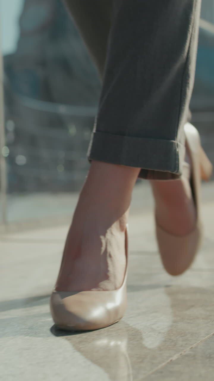 Cam bir binanın çevreyi yansıttığı ve yumuşak bokeh ışığıyla modern bir şehir yolunda yüksek topuklu ayakkabılarla kendinden emin bir şekilde yürüyen kızın düşük açıdan görünümü
