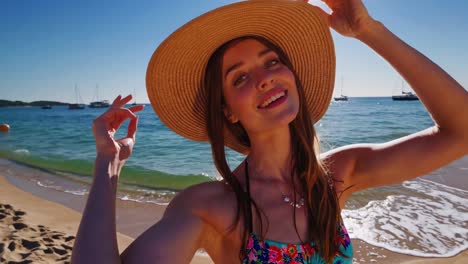 young woman wearing bikini and straw hat enjoying summer holidays on a sunny beach with sailboats on background, doing ok sign with hand
