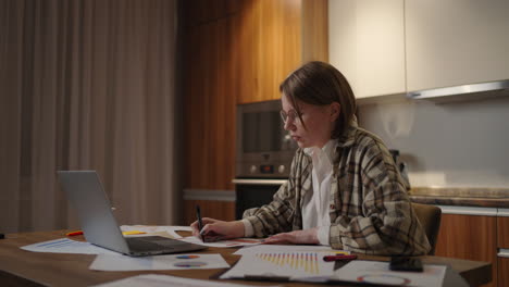 side view a woman with glasses works remotely from home sitting at a table with a laptop and a felt-tip pen marks the data on the graph. distance learning student at home will watch a lecture