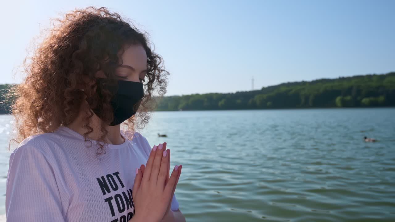 Jovem de cabelos cacheados meditando enquanto usa máscara de proteção preta. Parque e lago ao fundo. Dia ensolarado. Ideia sobre o coronavírus. Chisinau, Moldova