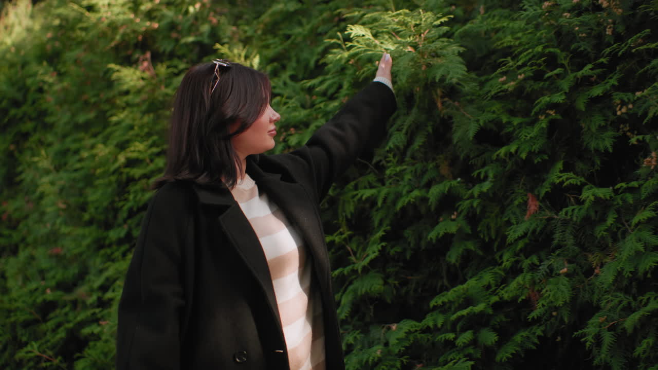 햇살 아래 긴 코트를 입고 머리에 선글라스를 얹은 세련된 보행자의 측면 모습이에요. 산책하는 동안 머리카락이 흩날리고 손은 공원 길을 따라 늘어선 녹색 울타리 옆 가을 단풍 위를 스치고, 잔잔한 바람이 불어요.