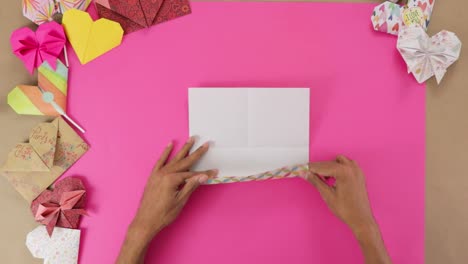 manos plegando papel origami sobre un fondo rosado vibrante rodeado de coloridos diseños de corazones, timelapse