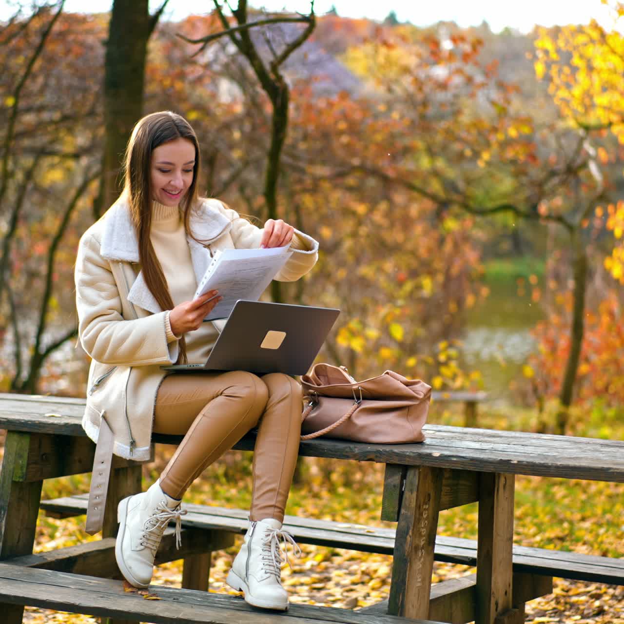 Smiling long-haired brunette lady looking through the papers in her hands. Woman sits on table in the park with laptop on her knees and having some remote work. Autmn nature at backdrop