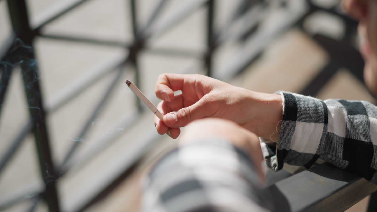 close up of smoker casually flicking lit cigarette as ash falls from tip in natural sunlight with soft blur background checkered shirt sleeve in frame