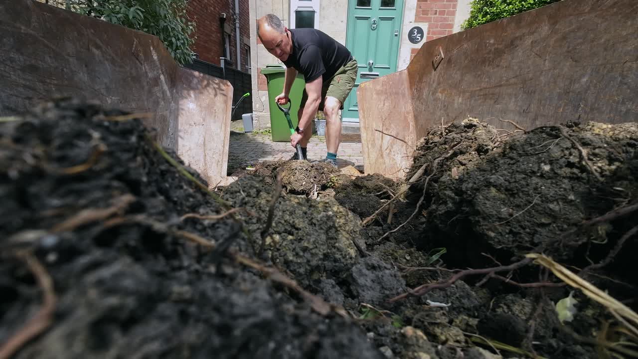 Wide shot from inside skip of middle aged man shovelling soil in to skip on a sunny day