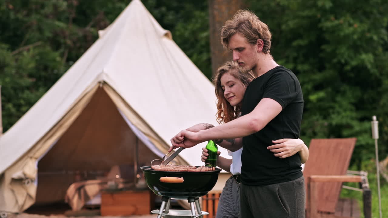 Young couple frying meat on the grill and drinking beer. Greenery around. Glamping. Slow motion