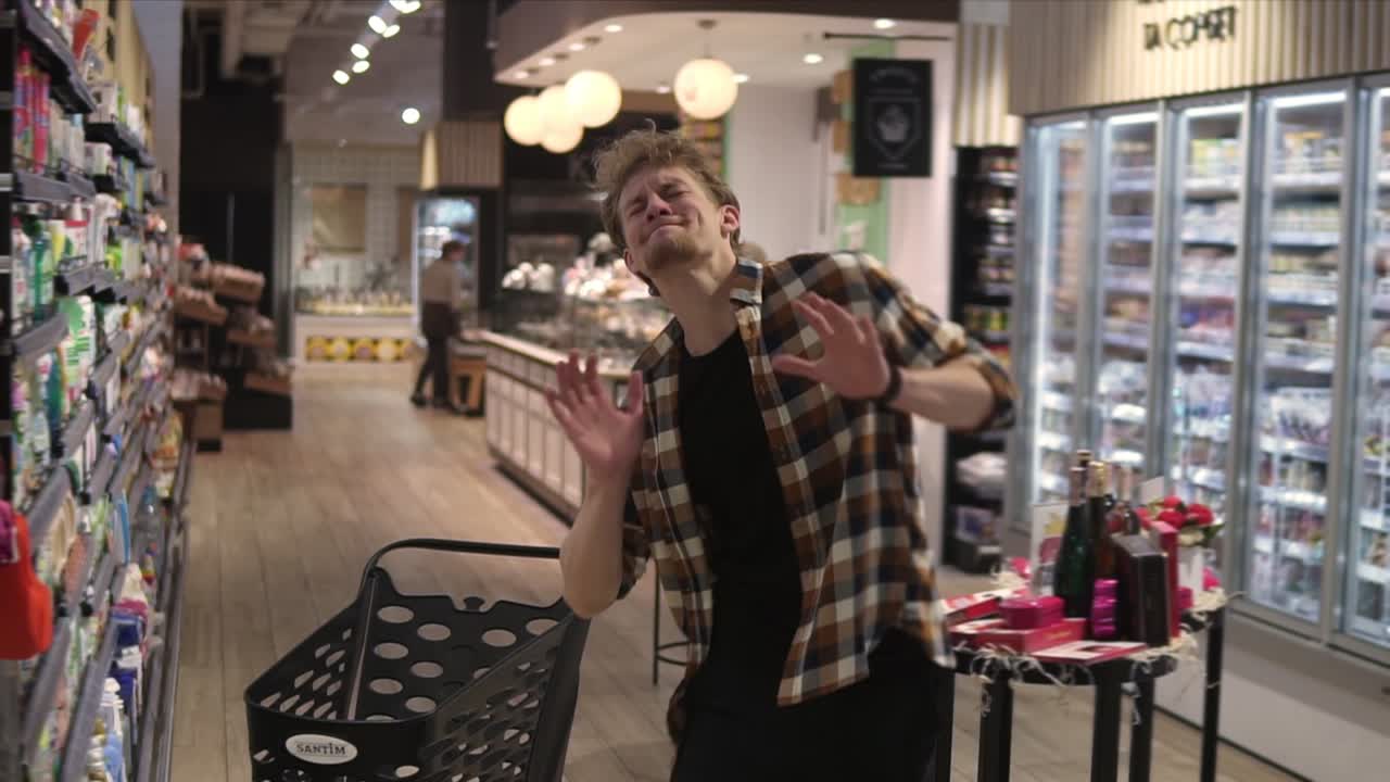 Excited young man in plaid shirt in supermarket dancing, funky moves close to shopping trolley. Positive dances in an empty food store row. Positive emotions