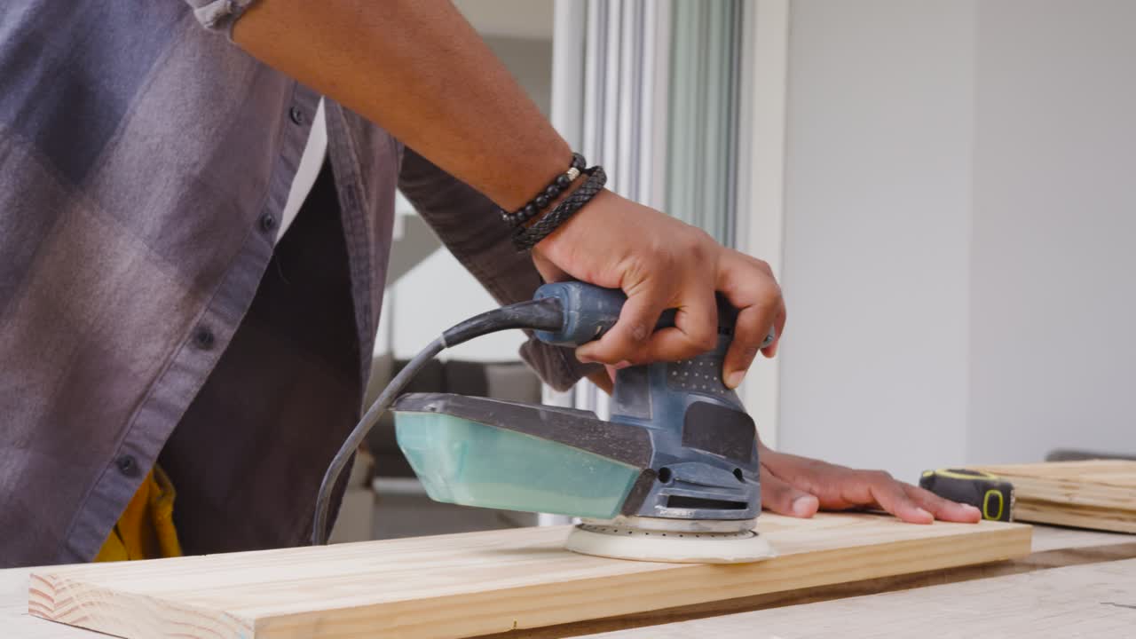 African American man powering corded sander guiding plank on home bench with goggles for finishing