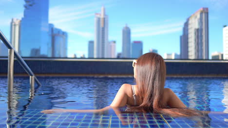 back view of brunette young woman leaning on the border inside rooftop swimming pool with bangkok skyscrapers and skyline in background