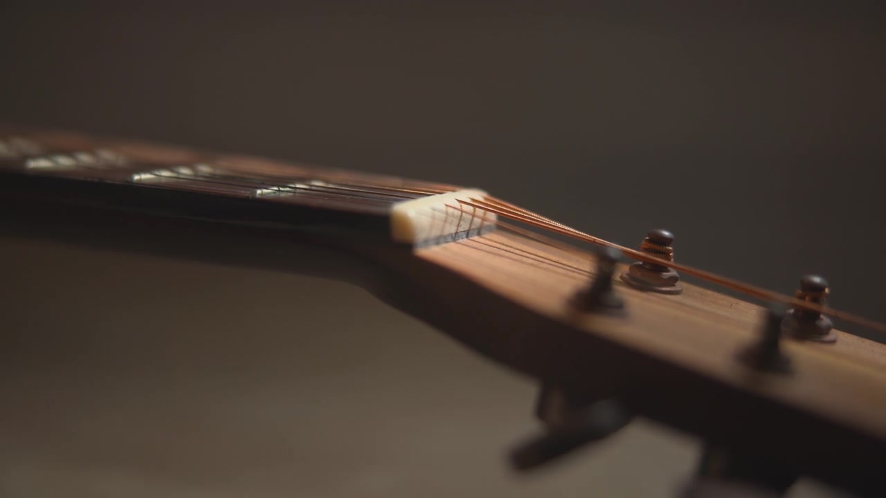 Vista ravvicinata della paletta di una chitarra acustica con corde tese e piroli di accordatura in metallo contro uno sfondo scuro e sfocato, illuminata da una luce calda e soffusa e con una profondità di campo ridotta.