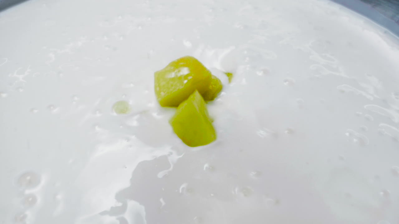 Slow motion close-up shot of mango cubes falling gently into a bowl of smooth white yogurt. The vibrant yellow contrasts with the creamy white texture, creating a visually delicious moment.