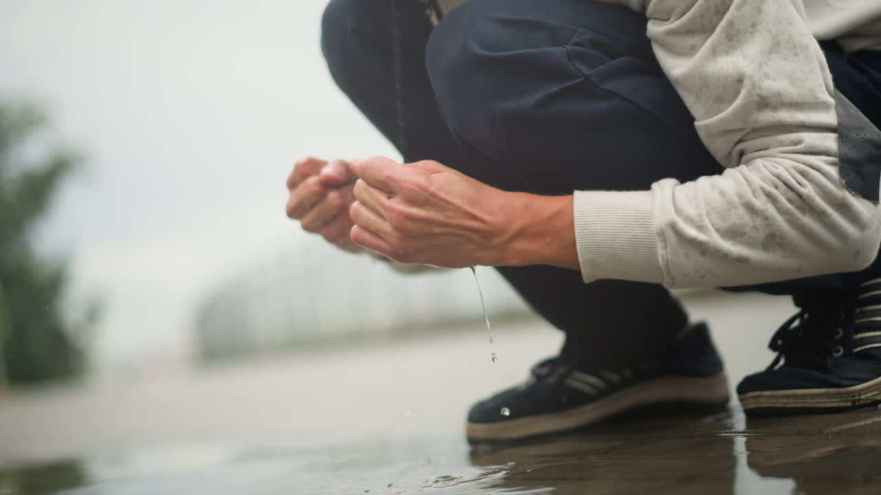 웅덩이 물을 퍼 올리는 손, 거리 탐험가가 반사된 물결로 놀고, 활발하게 물을 튀기고 공중으로 물방울을 흩뿌리며, 소매와 운동화 디테일이 보이는 웅크린 자세, 흐린 날씨의 인도가, 촉각적인
