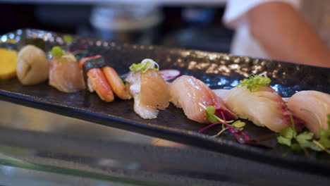 a colorful assortment of traditional nigiri sushi on black platter in japanese restaurant with chef in background, slider close up hd