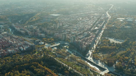 Aerial-panoramic-shot-of-river-flowing-through-city-with-parks.-Vehicles-driving-on-historic-Bridge-of-Segovia.-Town-illuminated-by-setting-sun.