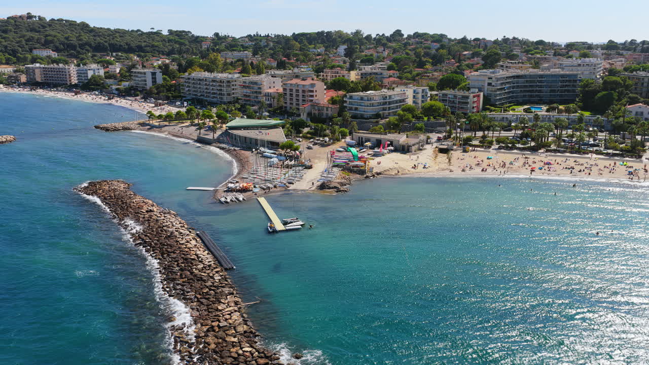 Aerial drone view of Antibes sandy beach with sunbathers, stone breakwaters, and sailing boats docked near the shoreline