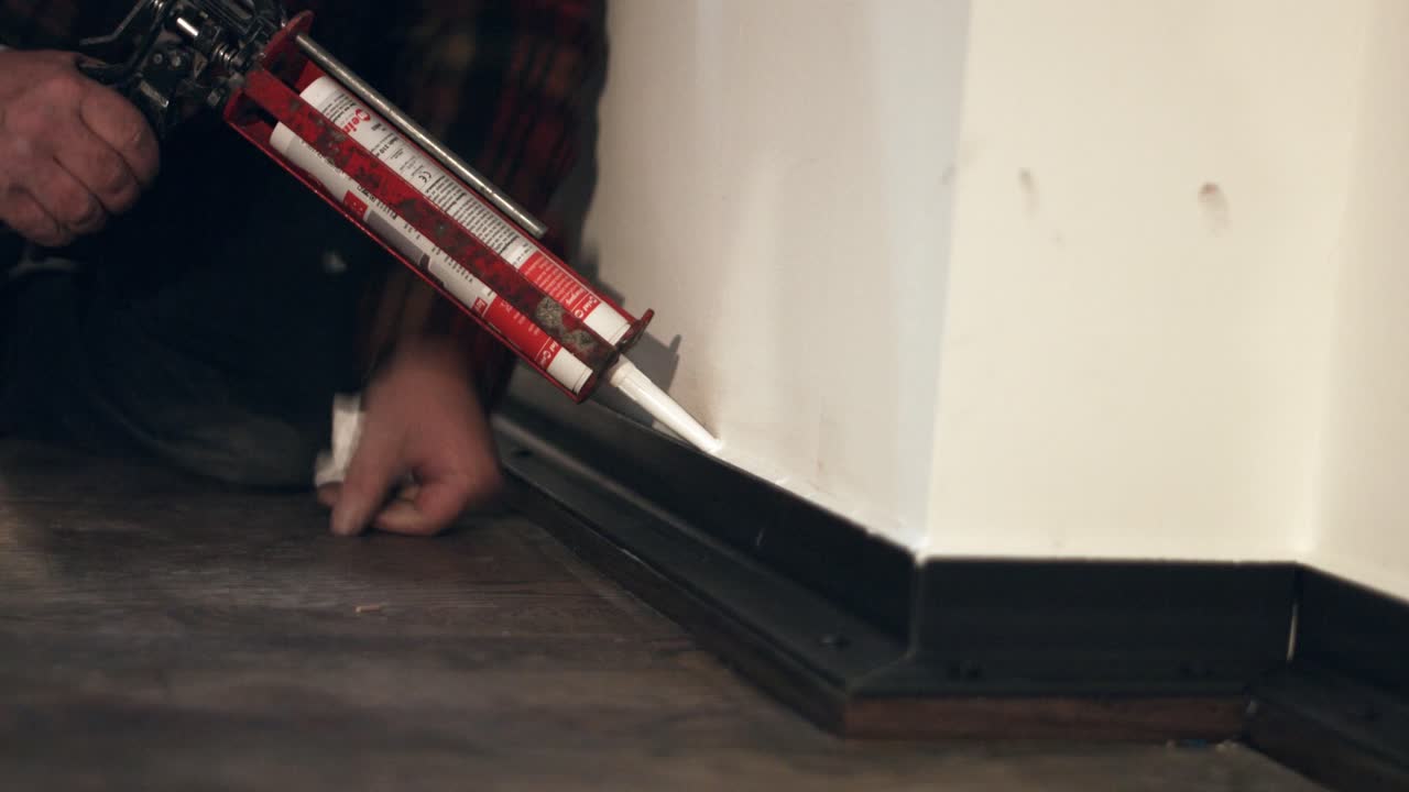Grout sealent out of silicon with a white color being applied on a skirting base board in a new apartment by a crafts person with a cartridge gun on a black wooden floor