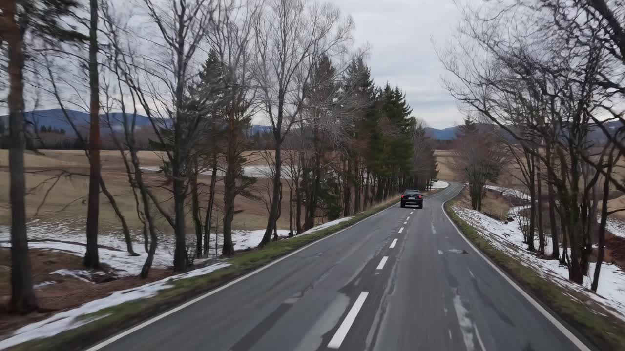Aerial video of a car driving on a winding road through a winter landscape, surrounded by bare trees