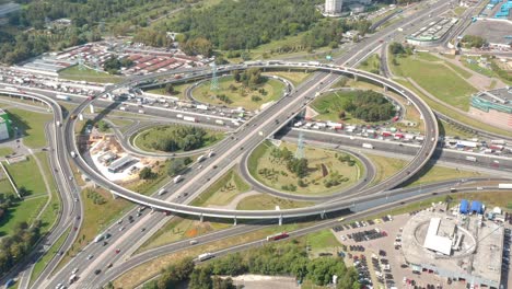 a large circular intersection with clover exits and ways. top aerial view from drone. everyday big city traffic. routine concept. sunny day. green areas with grass and buildings all around. camera is moving forward over the highway.