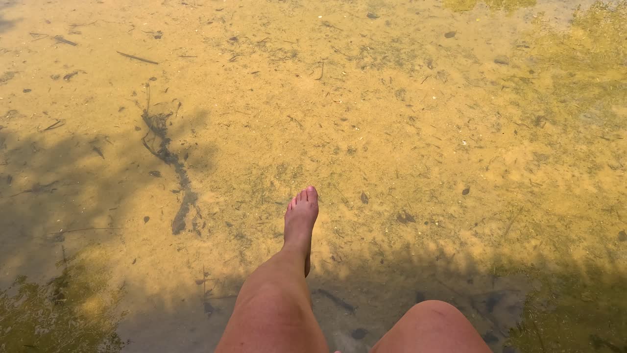 Bare legs gently swing above sunlit, shallow water with visible fish and sandy bottom. Natural daylight, steady overhead camera, tranquil outdoor setting