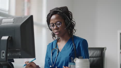 a black woman doctor wearing headphones sits at a table with a computer and takes calls from patients looks at their medical records and enters them into the clinic schedule