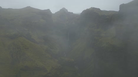 flying-through-clouds-to-reveal-a-majestic-waterfall,-canyon,-river,-and-green-valley