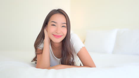 Attractive-Asian-woman-in-white-pajama-top-lying-on-large-soft-bed-with-white-linen