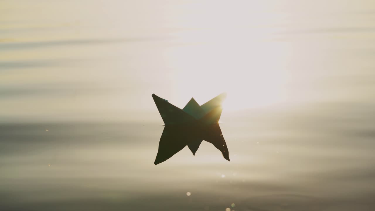 Paper boat floats alone with its reflection on the wavy water in the river at sunset. Beautiful rays of the evening sun falling on origami ship.Close-up