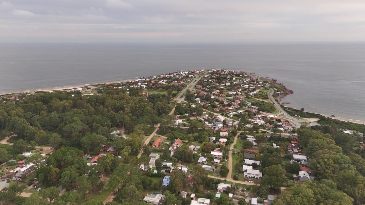 Punta Colorada si estende su una stretta penisola boscosa, con strade residenziali alberate e tetti circondati da un estuario calmo e dall'oceano sotto un cielo coperto. Maldonado, Uruguay. Ripresa aerea in sorvolo.
