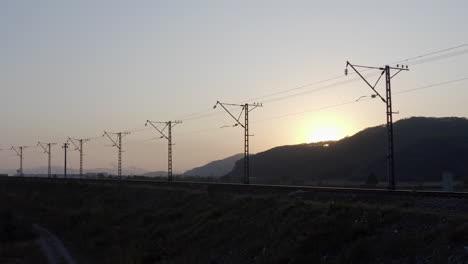 silueta de ferrocarril vacío con líneas eléctricas de alto voltaje y sol escondido detrás de una colina, al atardecer