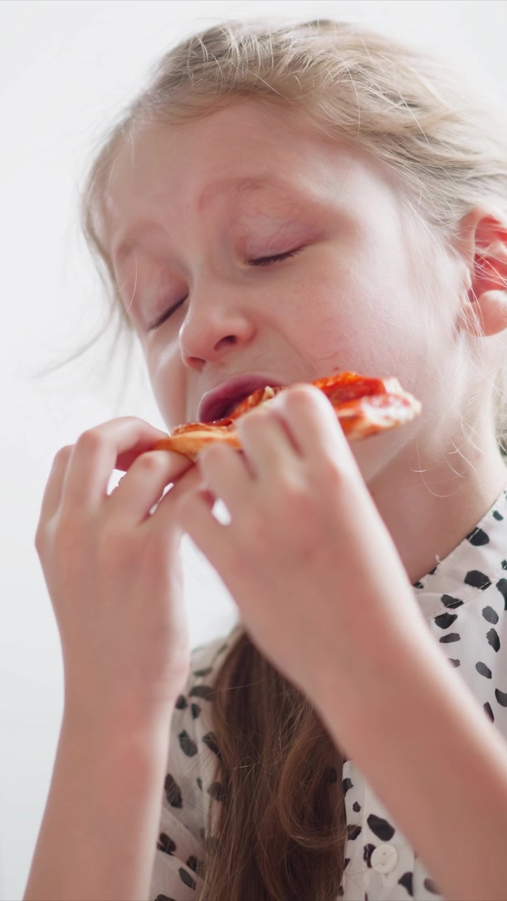 Close up of young girl eating small piece of food with both hands wearing white shirt with black spots. Soft lighting and plain background emphasize action without distractions