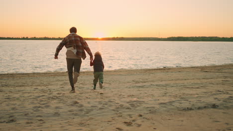 Fröhlicher-Vater-Und-Kleiner-Sohn-Ruhen-Sich-Aus-Und-Haben-Spaß-Am-Flussufer,-Der-Bei-Sonnenuntergang-Zum-Wasser-Rennt