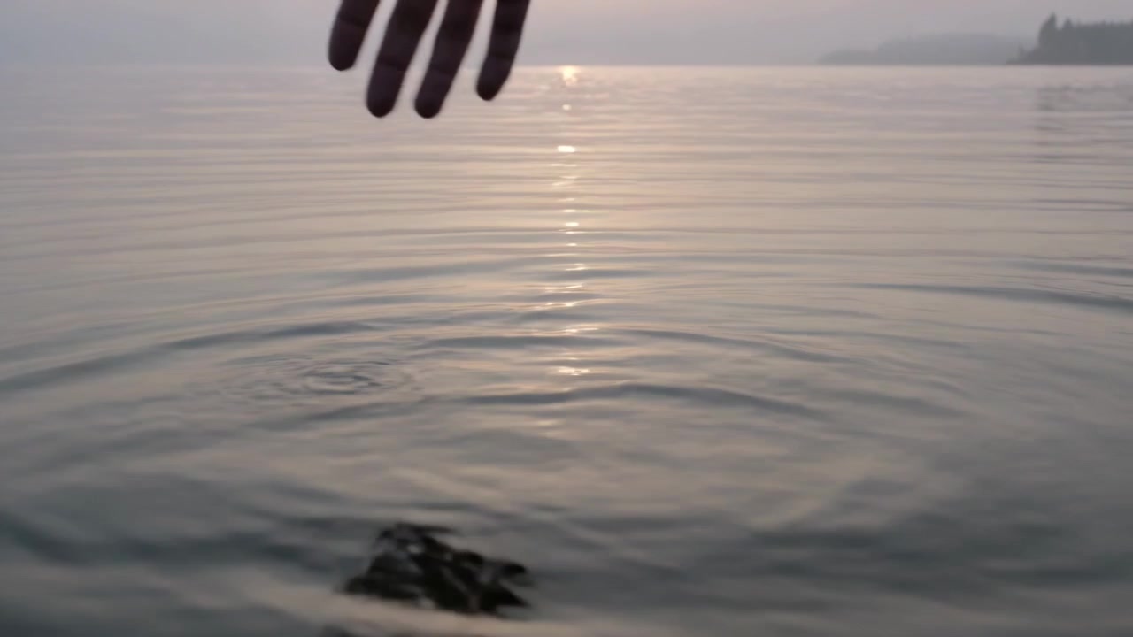 Mano nuda che scende creando increspature su un lago calmo con il sole basso, disturbando il riflesso.