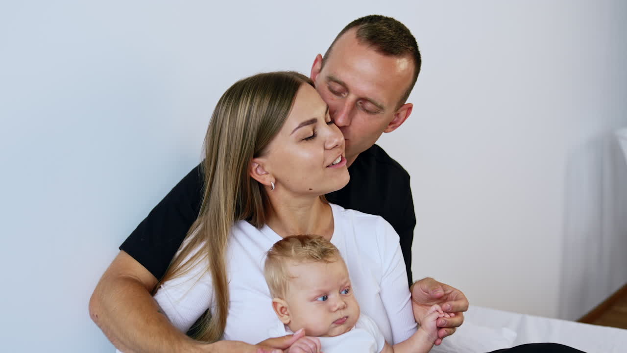 Husband kisses his wife holding a tiny blonde boy. Happy family of three at white backdrop.