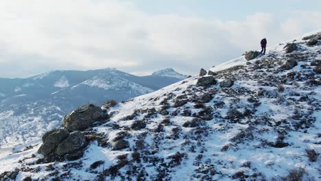 a traveler with a backpack climbs the slope of a hill covered with snow against the backdrop of a mountain panorama. top view with a circle flight