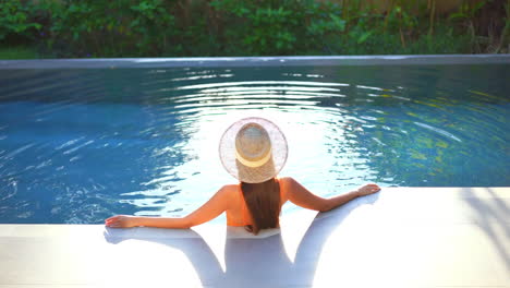 mujer elegante con un sombrero de pie en la piscina infinita disfrutando del paisaje tropical, concepto de moda de verano, fotograma completo estático