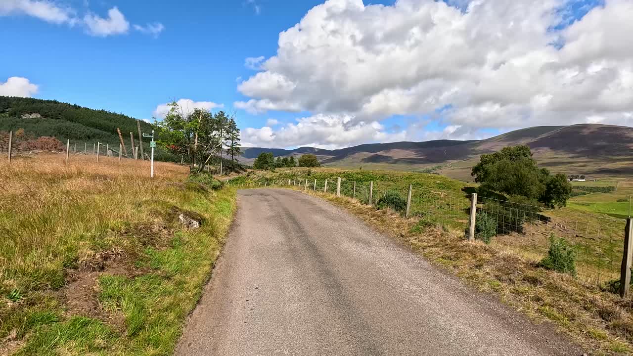 A vehicle travels a narrow, winding rural road through scenic Clova, Angus, Scotland, under bright daylight with blue skies and scattered clouds. Smooth camera movement captures the peaceful landscape