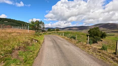 a vehicle travels a narrow, winding rural road through scenic clova, angus, scotland, under bright daylight with blue skies and scattered clouds. smooth camera movement captures the peaceful landscape