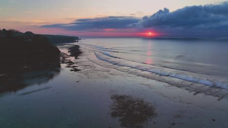 Drone-clip-showing-beautiful-pink-sunset-reflecting-on-open-calm-ocean-and-limestone-cliffs-of-Uluwatu,-Bali