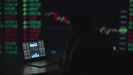 tarde en la noche en una oficina privada el hombre de negocios trabaja en una computadora portátil. tarde en la noche el comerciante lee números en sus múltiples pantallas con información de acciones en ellos