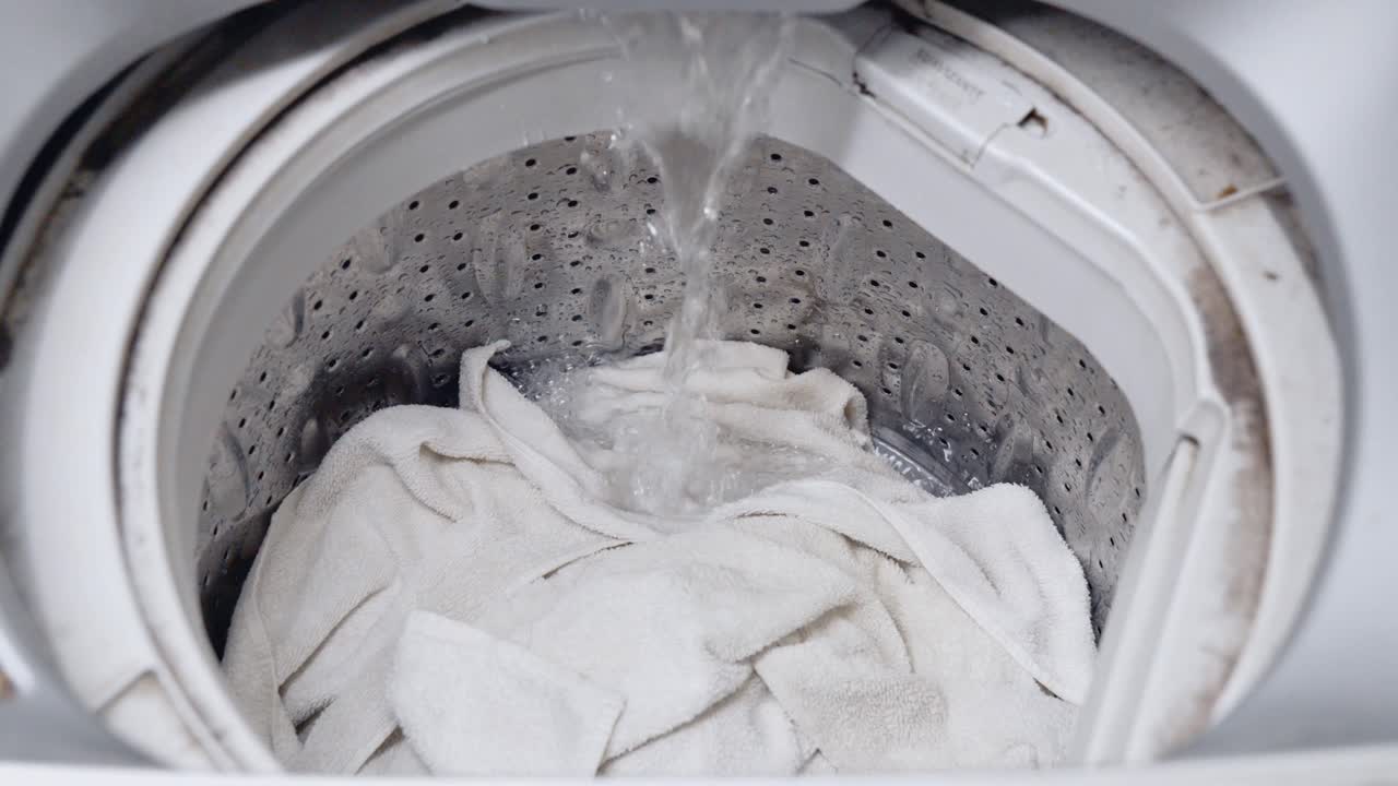 Clean water filling the stainless steel drum of a top-load washer with white towels. Laundry machine beginning a wash cycle at home