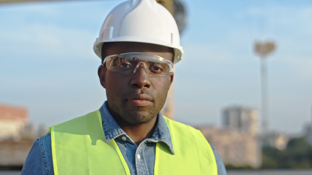 Close up of the African American young handsome man builder in hardhat and goggles looking at the camera and smiling. Outdoor at the building site.