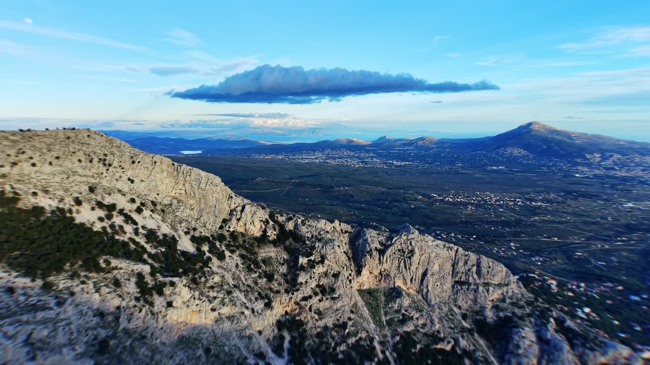 El dron se desliza hacia la izquierda desde la ladera de la montaña que alberga el Regency Casino Mont Parnes, enmarcando la cima rocosa. Más allá se extiende una vasta llanura que conduce a un mar lejano bajo un cielo azul.